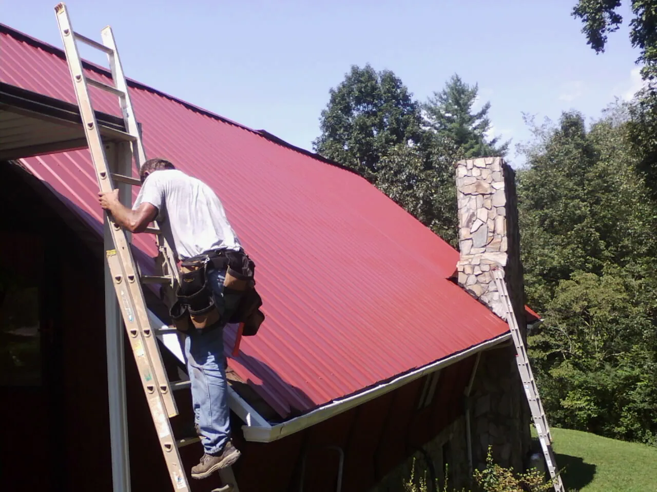 Roofer on a ladder beside a red metal roof and stone chimney — Soffit & Fascia Repair by Roofing Brothers in Devine